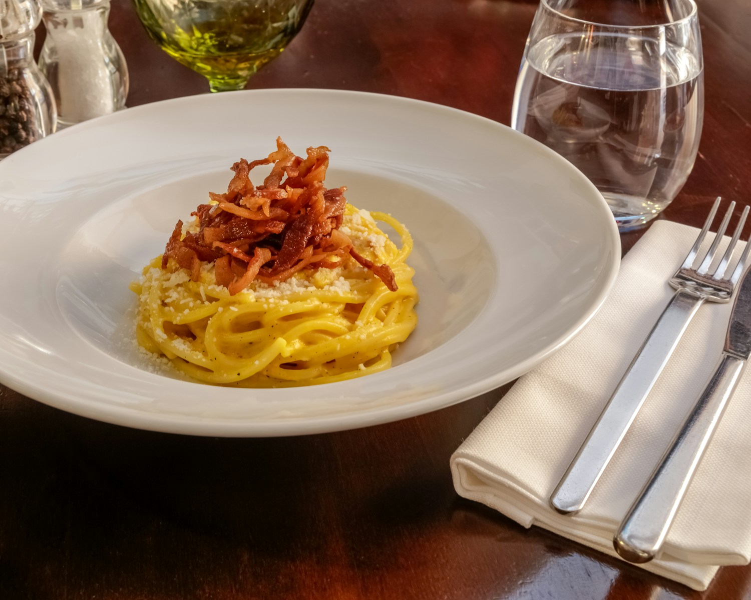 pasta with bacon on top on a round white ceramic plate near fork, knife, and glass of water